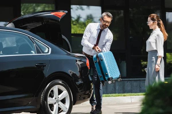 Selektiver Fokus einer lächelnden Geschäftsfrau, die neben einem Kollegen steht, der Koffer in den Kofferraum eines Autos auf der Stadtstraße legt — Stockfoto