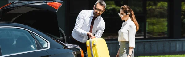 Panoramic shot of smiling businesswoman standing near colleague putting suitcase in car trunk outdoors — Stock Photo