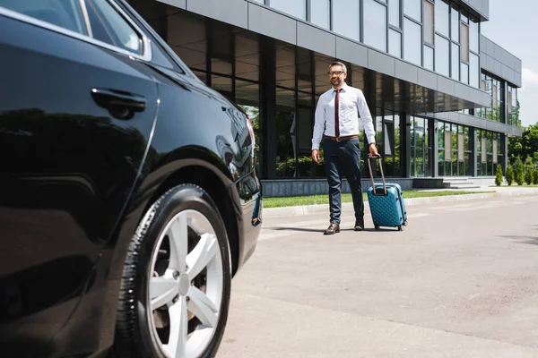 Selective focus of smiling businessman walking with suitcase to car on urban street — Stock Photo