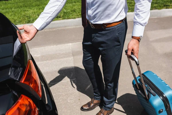Обрезанный вид бизнесмена, держащего чемодан и открывающего багажник автомобиля на городской улице — стоковое фото