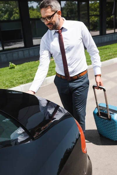 Handsome businessman holding suitcase and opening trunk of car on urban street — Stock Photo