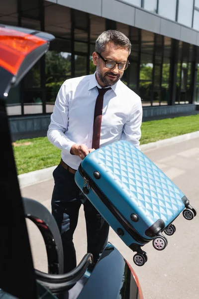 Selektiver Fokus eines gutaussehenden Geschäftsmannes mit Koffer in der Nähe von Auto mit offenem Kofferraum auf Stadtstraße — Stockfoto