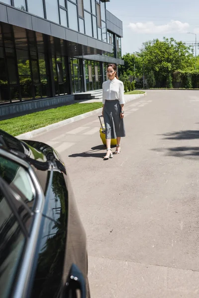 Focus selettivo di attraente donna d'affari che tiene valigia e cammina verso l'auto sulla strada urbana — Foto stock