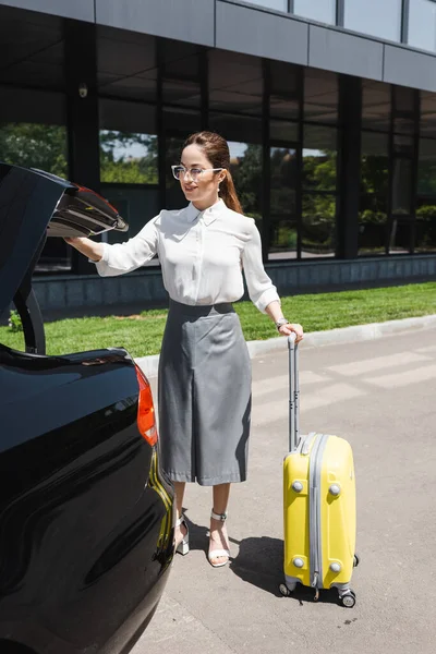 Hermosa mujer de negocios sonriente sosteniendo la maleta cerca del coche con baúl abierto en la calle urbana - foto de stock