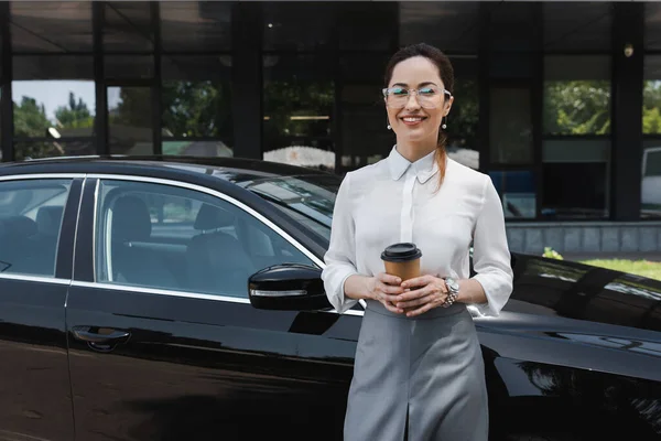 Красивая деловая женщина улыбается перед камерой, держа в руках бумажную чашку рядом с автомобилем на городской улице — стоковое фото