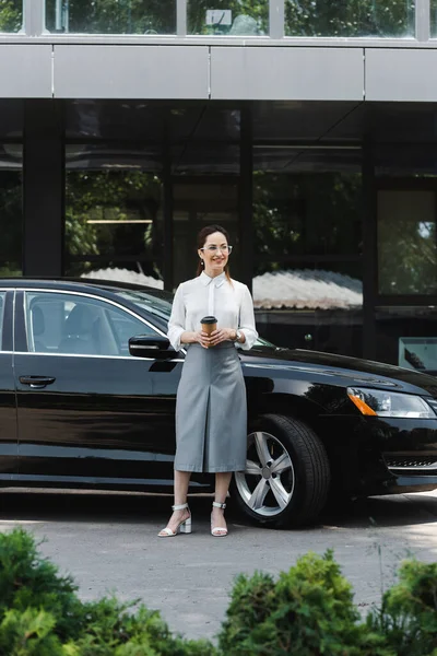 Focus selettivo di donna d'affari sorridente che tiene il caffè per andare vicino auto sulla strada urbana — Foto stock