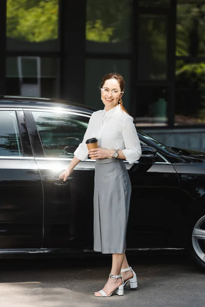Empresária sorridente segurando copo de papel e abrindo a porta do carro na rua urbana — Fotografia de Stock