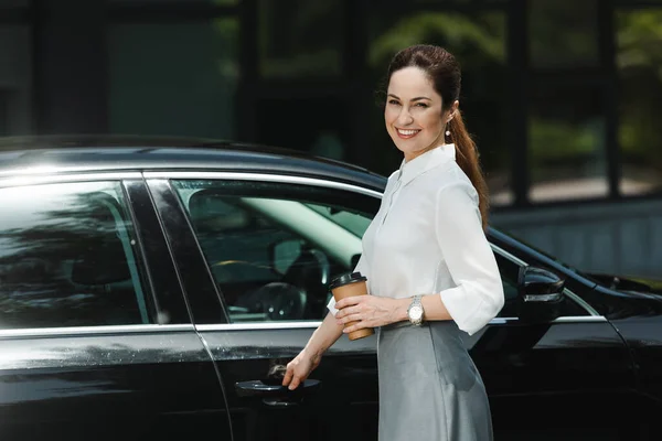 Side view of beautiful businesswoman smiling at camera while holding paper cup and opening car door on urban street — Stock Photo