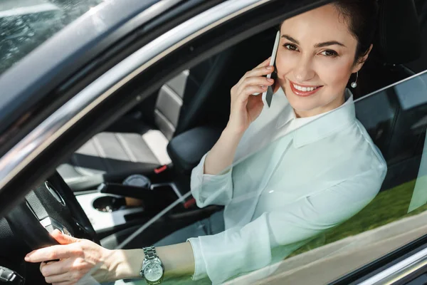 Foco seletivo de empresária sorrindo para a câmera e falando no smartphone no assento do motorista em auto — Fotografia de Stock