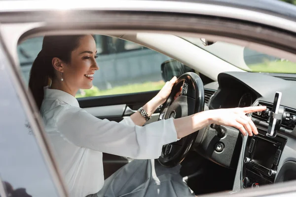 Vista lateral de una mujer de negocios sonriente usando un teléfono inteligente mientras conduce auto - foto de stock