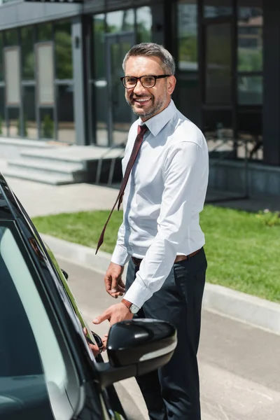 Selective focus of businessman smiling at camera and opening door of car on urban street — Stock Photo