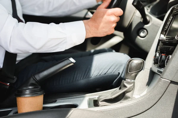 Vista recortada del hombre de negocios en coche de conducción de desgaste formal cerca de la taza de papel en portavasos a la luz del día — Stock Photo