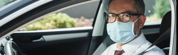 Панорамна орієнтація бізнесмена в медичній масці, дивлячись на камеру на сидінні водія в машині — стокове фото