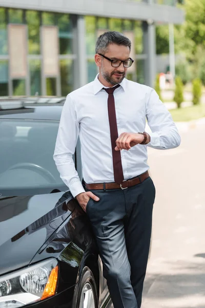 Hombre de negocios guapo mirando un reloj de pulsera cerca del coche en la calle urbana - foto de stock