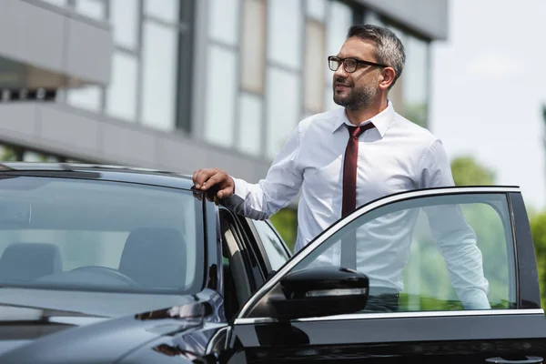 Вибірковий фокус красивого бізнесмена, що стоїть біля автомобіля з відкритими дверима на міській вулиці — стокове фото