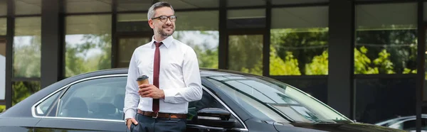 Vue panoramique d'un homme d'affaires souriant tenant une tasse en papier près d'une voiture dans une rue urbaine — Photo de stock