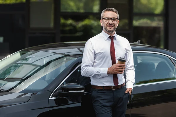 Hombre de negocios guapo sonriendo a la cámara y sosteniendo café para ir cerca de auto en la calle urbana — Stock Photo