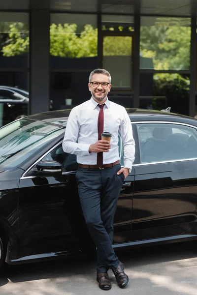 Homme d'affaires prospère souriant à la caméra tout en tenant une tasse de café près de la voiture dans la rue urbaine — Photo de stock