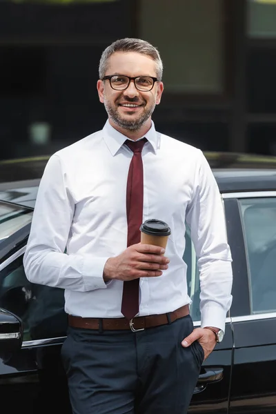 Красивый бизнесмен держит кофе, чтобы подойти к машине и улыбается в камеру на городской улице — стоковое фото