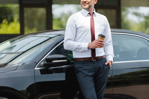 Vista ritagliata dell'uomo d'affari sorridente che tiene la tazza di carta vicino all'auto sulla strada urbana — Foto stock