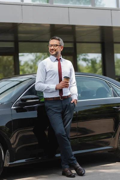 Homme d'affaires positif regardant loin tout en tenant du café pour aller près de l'auto sur la rue urbaine — Photo de stock