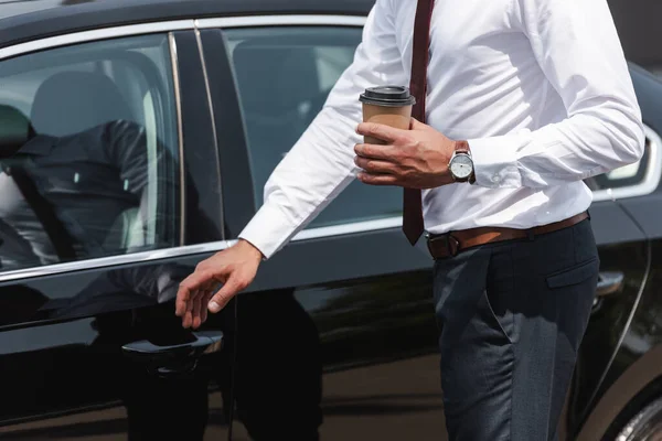 Vista recortada del hombre de negocios sosteniendo café para ir y abrir la puerta del coche en la calle urbana — Stock Photo