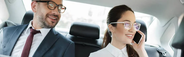 Panoramic crop of smiling businesswoman talking on smartphone near businessman in auto — Stock Photo