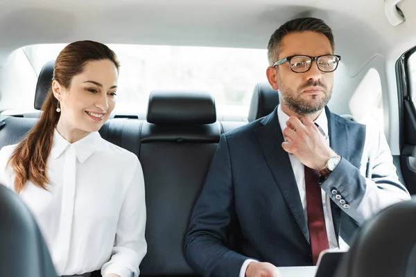 Selective focus of smiling businesswoman sitting near businessman using laptop in auto — Stock Photo