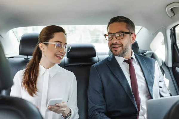 Selective focus of smiling business people using smartphone and laptop in car — Stock Photo