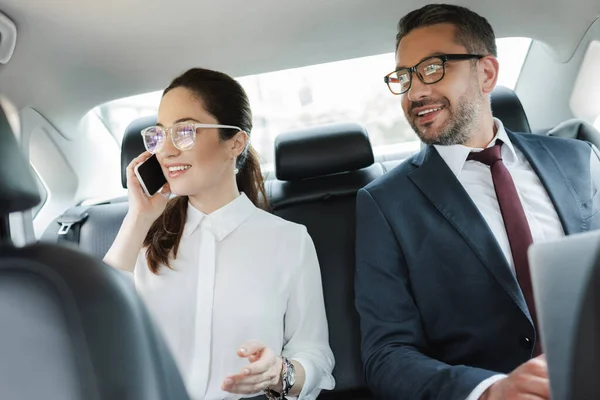 Foco seletivo de empresária positiva falando no smartphone perto de empresário com laptop no carro — Fotografia de Stock