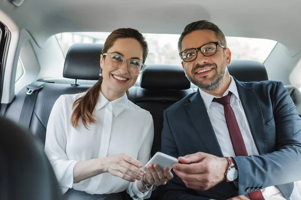 Enfoque selectivo de la mujer de negocios sonriente sosteniendo el teléfono inteligente y mirando a la cámara cerca de hombre de negocios en coche - foto de stock