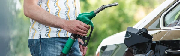 Panoramic crop of man holding fueling nozzle near open gas tank cover of car on urban street — Stock Photo