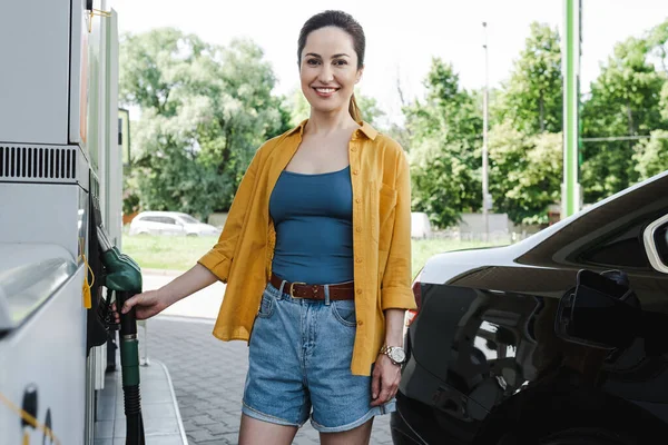 Enfoque selectivo de la hermosa mujer sonriente sosteniendo la boquilla de repostaje cerca del coche en la gasolinera - foto de stock