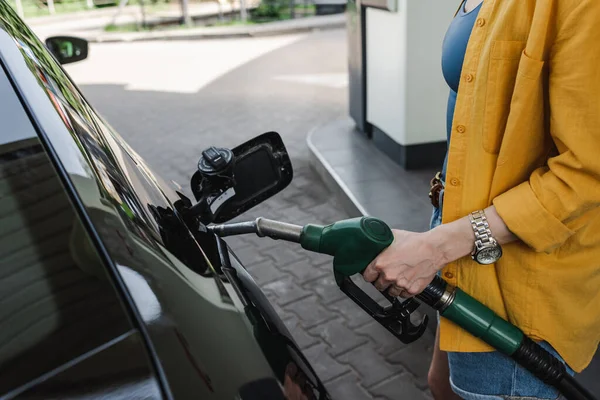 Vista recortada de la mujer sosteniendo la boquilla de carga cerca del tanque de gasolina del coche en la calle urbana - foto de stock