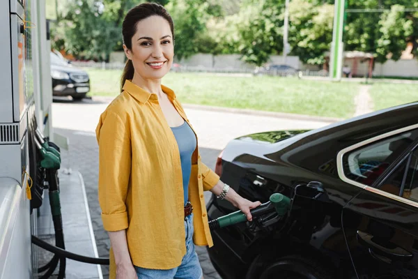 Selektiver Fokus einer Frau, die in die Kamera lächelt, während sie Auto an Tankstelle tankt — Stockfoto