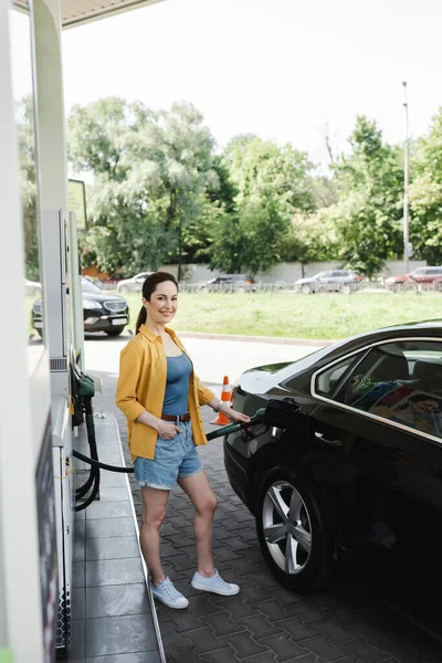 Selective focus of smiling woman with hand in pocket looking at camera and fueling car on gas station — Stock Photo