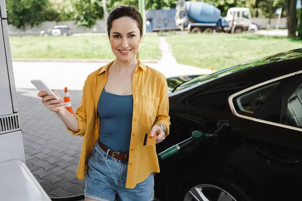 Mulher sorridente segurando cartão de crédito e smartphone perto do carro no posto de gasolina — Fotografia de Stock