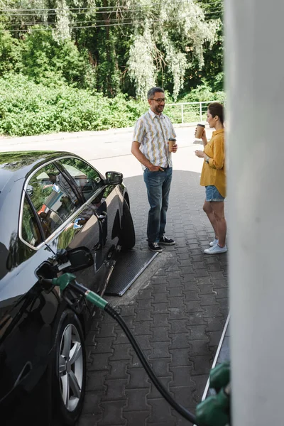 Selektiver Fokus eines lächelnden Mannes, der Kaffee hält, um sich seiner Frau zu nähern, während er Auto an Tankstelle tankt — Stockfoto