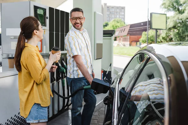 Selective focus of smiling man looking at wife with coffee to go while refueling car on gas station — Stock Photo