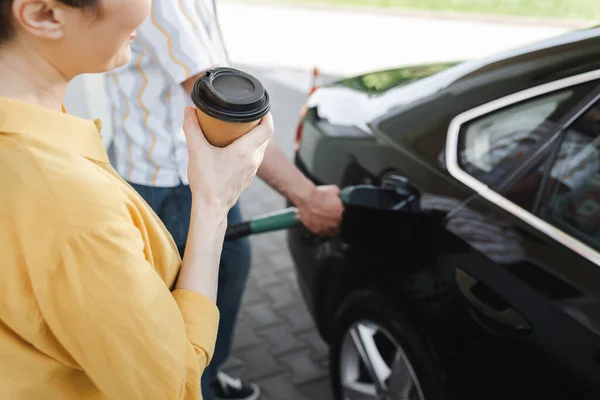 Vista cortada de mulher segurando café para ir enquanto marido alimentando auto no posto de gasolina — Fotografia de Stock