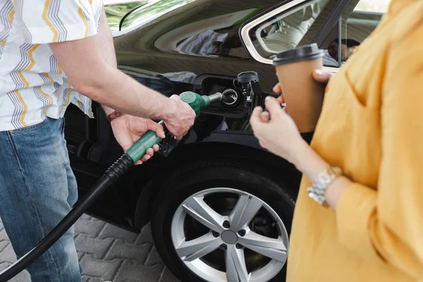 Vista ritagliata dell'uomo che tiene l'ugello di rifornimento vicino al serbatoio di gas dell'auto vicino alla donna con tazza di carta sul distributore di benzina — Foto stock