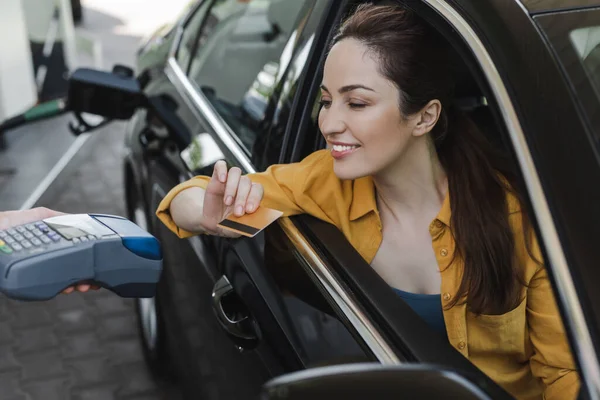 Foco seletivo de mulher sorridente que mantém o crédito no cartão perto do funcionário do posto de gasolina com terminal de pagamento — Fotografia de Stock