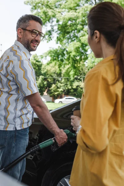 Вибірковий фокус усміхненого чоловіка, який заправляє машину і дивиться на дружину з паперовою чашкою на автозаправці — стокове фото