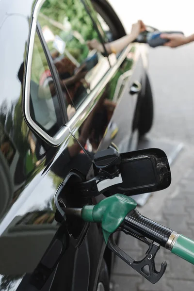 Foco seletivo do bico de abastecimento no tanque de gás do carro e da mulher pagando com cartão de crédito no posto de gasolina — Fotografia de Stock