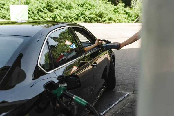 Mitarbeiter einer Tankstelle mit Zahlungsterminal in der Nähe von Frau mit Kreditkarte im Auto im Fokus — Stockfoto