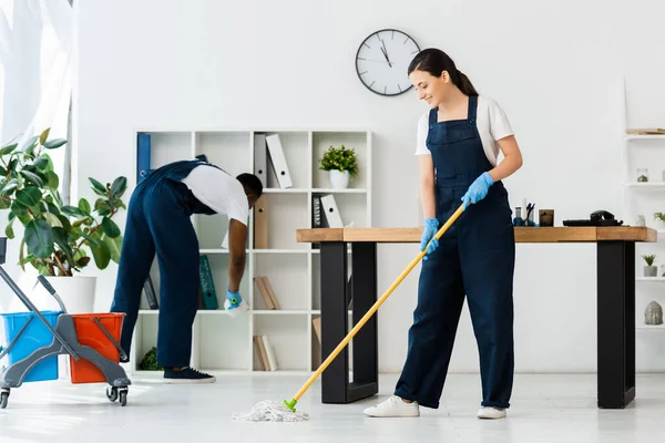 Focus sélectif de nettoyage souriant plancher de lavage avec serpillière près d'un collègue afro-américain au bureau — Photo de stock