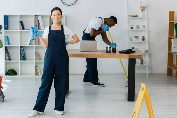 Focus selettivo del sorridente pulitore tenendo straccio vicino al collega afro-americano che lava il pavimento in ufficio — Foto stock