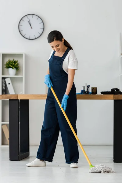 Nettoyant souriant dans des gants en caoutchouc plancher de lavage avec serpillière dans le bureau — Photo de stock