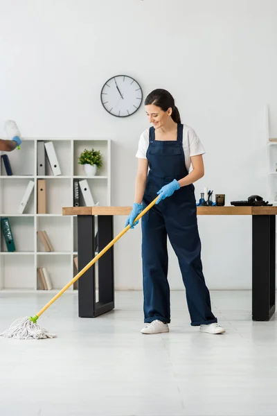 Lächelnder sauberer Waschboden mit Wischmopp in der Nähe eines amerikanischen Kollegen im Büro — Stockfoto