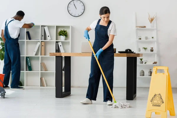 Foco selectivo del letrero de suelo húmedo y limpiadores multiétnicos que trabajan en la oficina - foto de stock